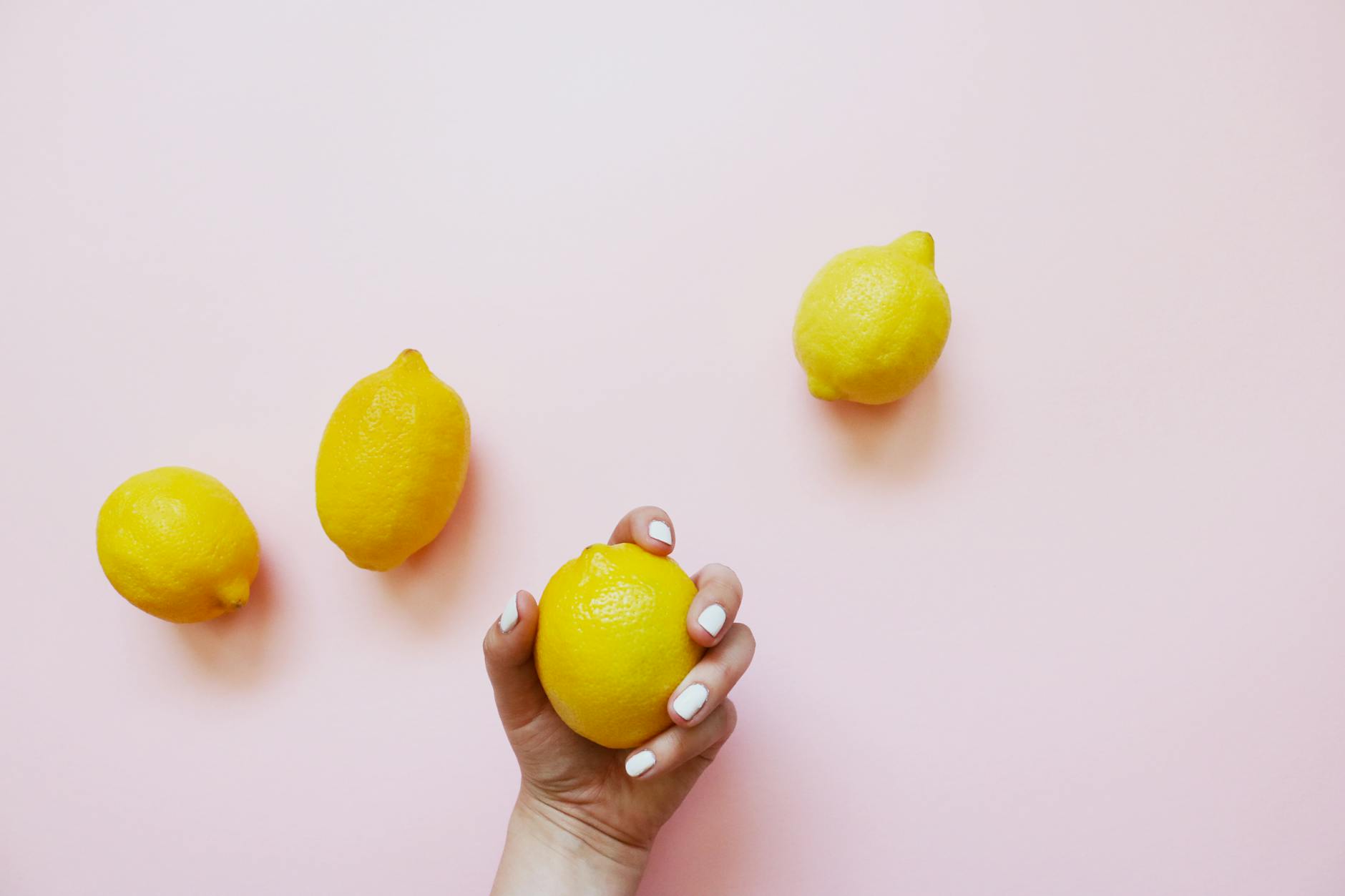A hand holding a yellow lemon against a soft pink background surrounded by fresh lemons