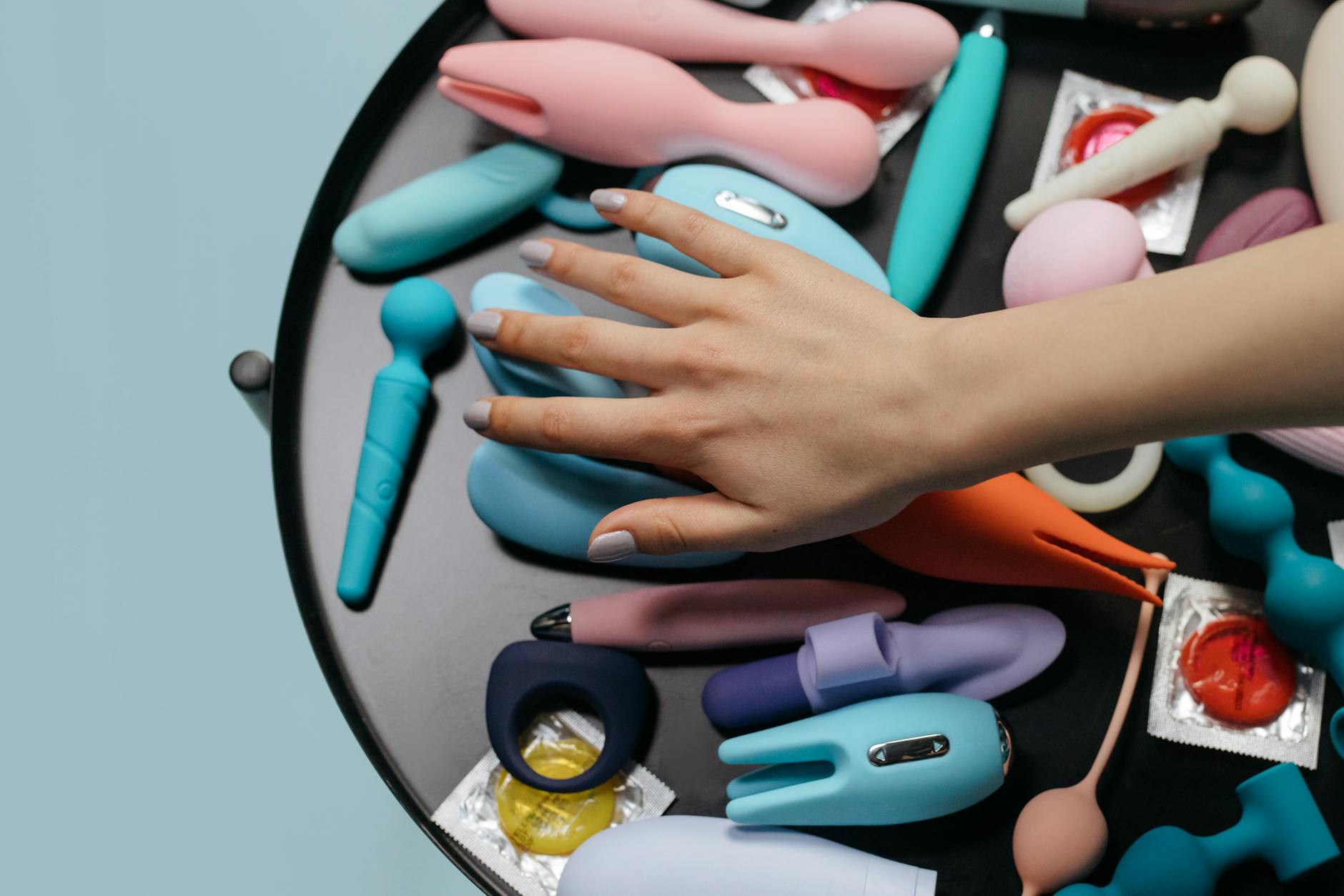 A hand reaching over a variety of colorful clitoral vibrators and sex toys arranged on a table.
