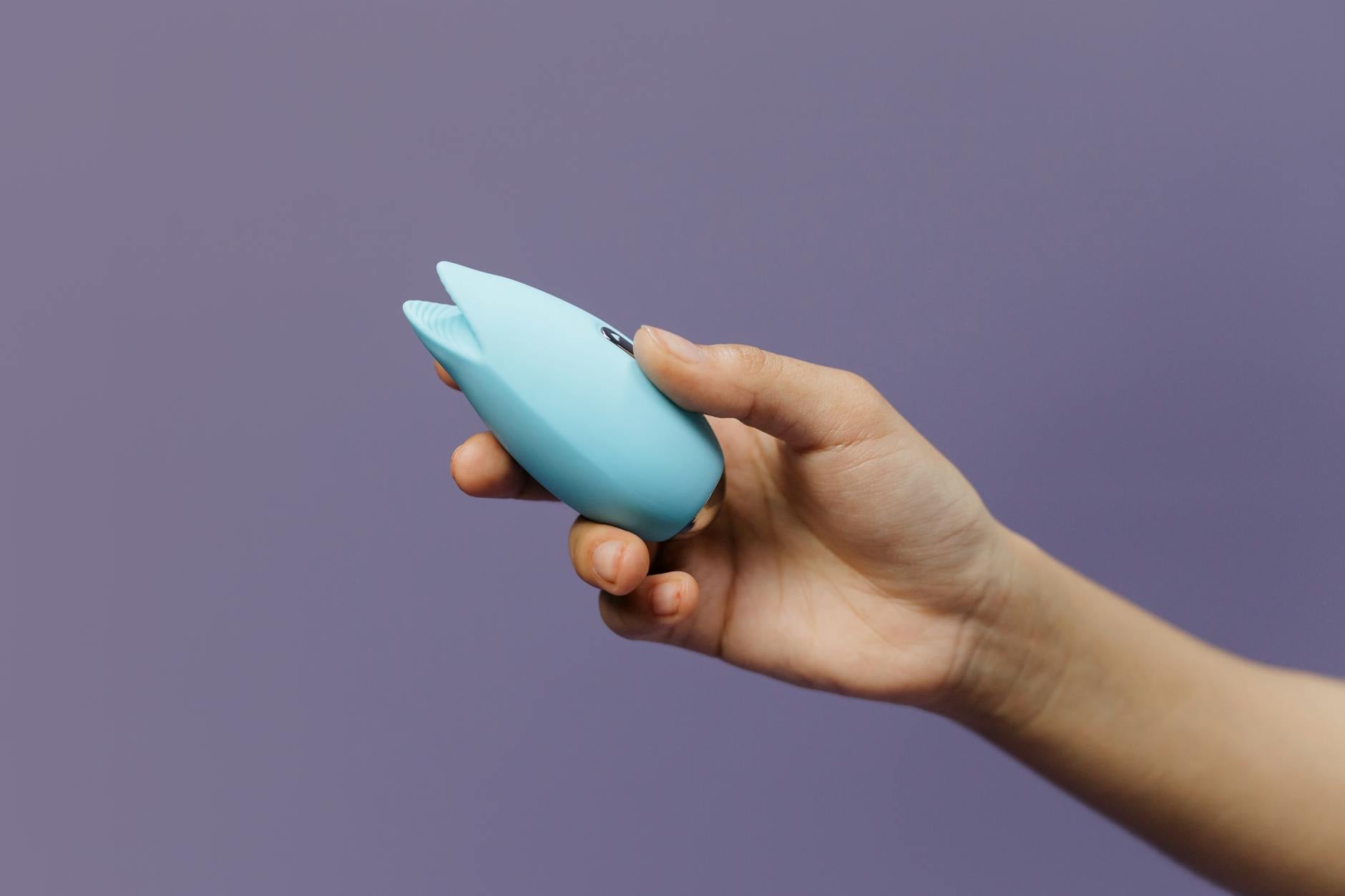 A blue silicone clitoral vibrator held against a purple background, representing pleasure devices designed for sensitive tissue.