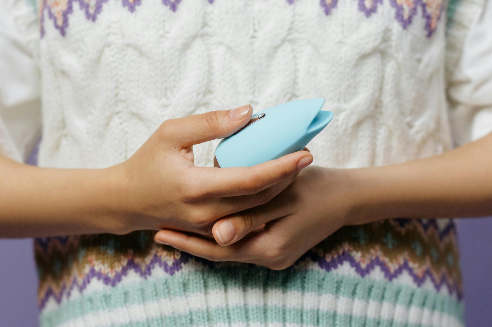 Primer plano de manos sosteniendo un vibrador de color azul contra un suéter tejido.